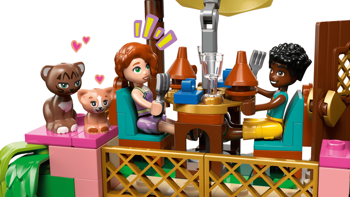 eine Szene in einem Cafe. Alles aus Legosteinen. Rechts und links von einem braunem Tisch sind zwei braun, türkise Stühle. Links sitzt eine Legofigur, die eine lila Hose, die auf den Oberschenkeln endet und ein lila Top trägt. Die Figur hat rote lange Haare, geschminkte grüne Augen und eine helle Hautfarbe. Auf dem rechten Platz sitzt eine Figur mit schwarzer Hautfarbe schwarzen kurzen Haaren, einem türkisen kurzärmligenT Shirt, und einer gelben Hose, die bis zu den Unterschenkel reicht. links neben dem Cafe sitzen 2 Katzen, eine dunkelbraune größere und eine hellbraune kleinere, um die kleinen, pinken Herzen fliegen.