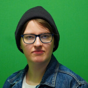 Ein Porträt von Team-Mitglied Samu. Zu sehen ist eine weiße Person mit dunkelblonden Haaren. Dey trägt eine schwarze Mütze, eine Brille und eine Jeansjacke über einem weißen Shirt. Der Hintergrund ist einfarbig grün.