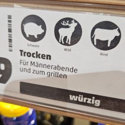 Foto von Schild am Weinregal oben sind 3 runde, schwarze Kreise mit einem Schwein, Hirsch und einem Rind Darunter geschrieben: Trocken Für Männerabende und zum Grillen