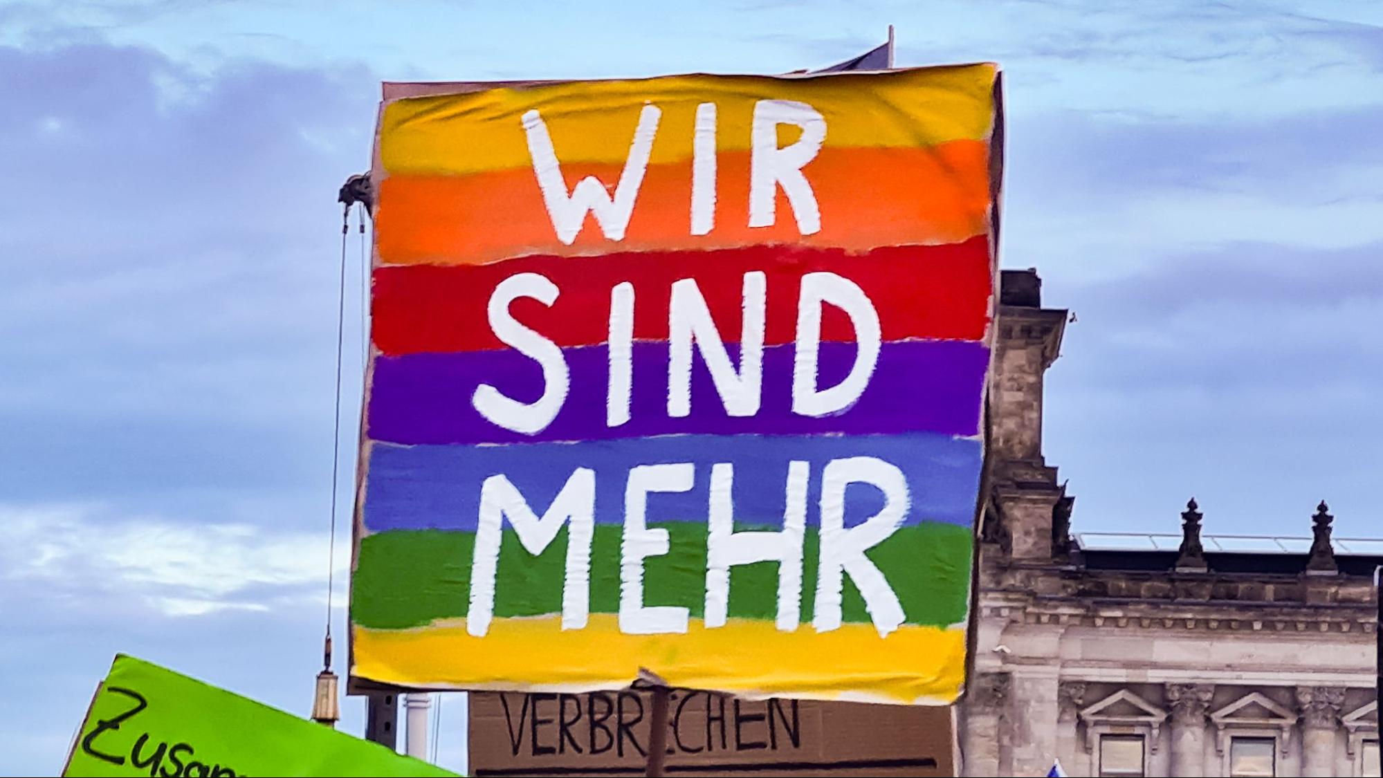 Foto von einem Demoplakat. Das Plakat ist in den Farben des Regenbogens angemalt und darauf steht in weiß WIR SIND MEHR