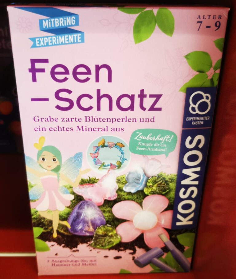 Feen-Schatz: eine rosa Kartonverpackung mit der Aufschrift “Mitbring Experiment Feen-Schatz grabe zarte Blütenperlen und ein echtes Mineral aus” darunter zu sehen eine gezeichnete Fee und rosa und lilafarbene Steinchen in Form von Schmetterling und Blumen.