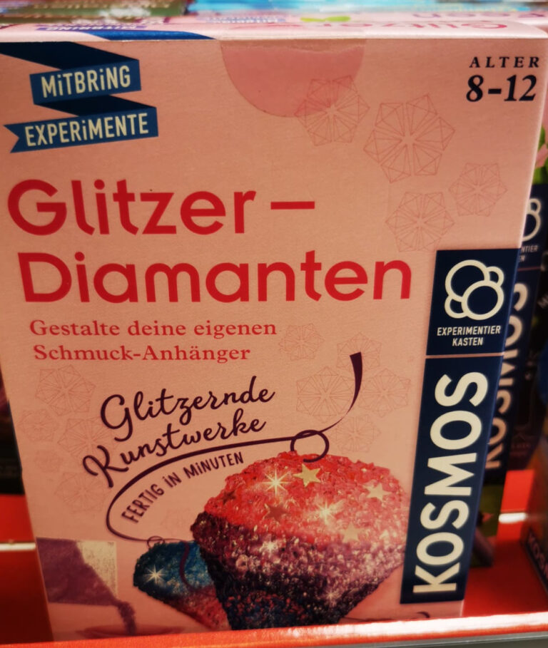 Glitzer-Diamanten: eine rosa Kartonverpackung mit der Aufschrift “Mitbring Experimente Glitzer-Diamanten Gestalte deinen eigenen Schmuckanhänger” darunter 2 rosalilafarbene Glitzersteine