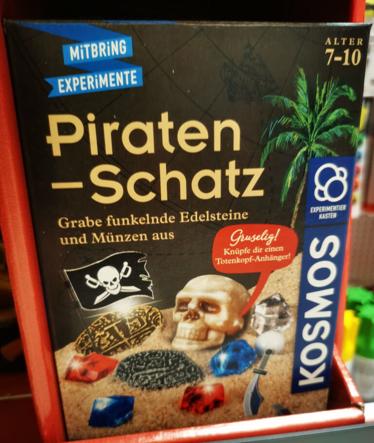 Piratenschatz:eine graue Kartonverpackung mit der Aufschrift “Mitbring Experiment Piraten-Schatz Grabe funkelnde Edelsteine und Münzen aus” darunter ist eine Schicht Sand, auf der Spielzeugedelsteine, alte Münzen und ein SPielzeugschädel liegt.