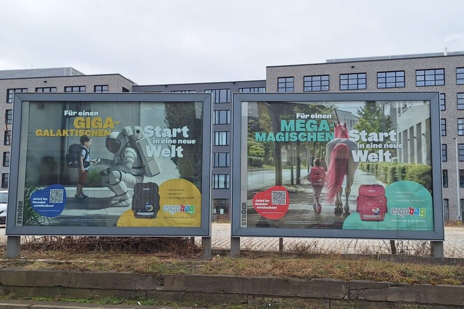 Zwei große Werbeplakate von ergobag. Auf einem ein Junge mit blauem Ranzen, welcher einer/einem Astronaten/ Astronautin die Hand gibt. Auf dem anderen Plakat ein Mädchen, welches mit einem pinken Ranzen neben einem Einhorn läuft.