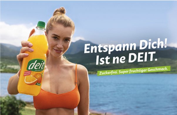 Werbeanzeige für DEIT-Limo mit einer jungen, blonden Frau im orangefarbenen Bikini-Oberteil, die eine gelbe Flasche des beworbenen Produkts in die Kamera hält. Sie steht vor einem blauen Badesee mit Bergen im Hintergrund. Ihr Haar ist zu einem hohen Dutt gebunden und sie lächelt von unten herauf in die Kamera. Der Werbetext lautet: "Entspann Dich! Ist ne DEIT." mit dem Zusatz "Zuckerfrei. Super fruchtiger Geschmack." in einem grünen Banner.