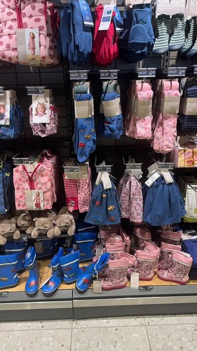 Foto eines Verkaufregals mit Kinderkleidung. Zu sehen sind Mützen, Strumpfhosen und Gummistiefel. Ebenfalls die Auswahl zwischen rosa mit Blümchen und blau mit Autos.