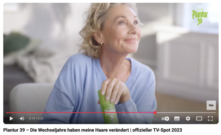 Screenshot eines YouTube-Videos einer Werbung für Plantur39- Haarpflegeprodukte. Das Bild zeigt eine Frau mittleren Alters mit grau-blonden, welligen Haaren in einem hellblauen Pullover. Sie sitzt entspannt in einem hellen, modern eingerichteten Wohnzimmer, Sonnenlicht fällt auf ihr Gesicht und sie hält eine grüne Plantur39-Flasche in der Hand. Ihr Gesichtsausdruck ist zufrieden, sie lächelt glücklich, während sie nach oben blickt. Der Titel des Videos lautet "Plantur 39 – Die Wechseljahre haben meine Haare verändert | offizieller TV-Spot 2023”.