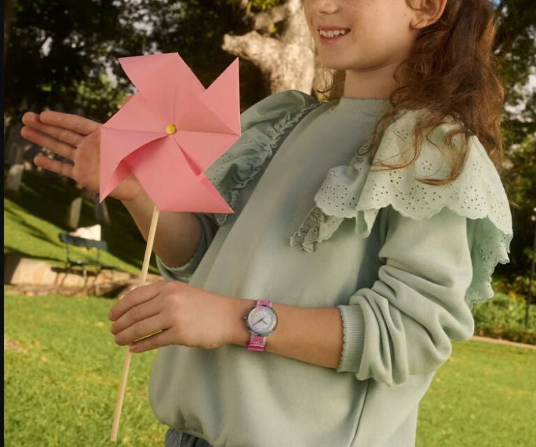 zu sehen ist der Oberkörper eines Mädchen mit langen, braunen Haaren und einem grünen Oberteils mit Rüschchen. Das Kind trägt eine rosafarbene Uhr und hält in der linken Hand ein rosa Windrad.