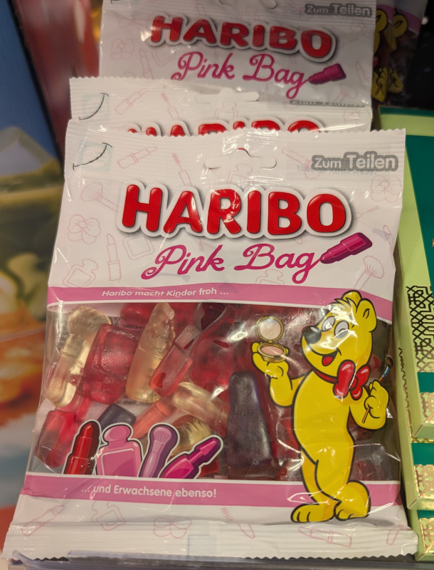 Mehrere Packungen „HARIBO Pink Bag“ stehen in einem Verkaufsregal. Die weiße Verpackung ist mit rosa Schrift und Illustrationen von Kosmetikartikeln gestaltet. Durch das Sichtfenster sind rote, rosa, lilafarbene und helle Fruchtgummis zu sehen. Auf der Packung hält der gelbe HARIBO-Goldbär einen Taschenspiegel und trägt eine rote Schleife. Daneben sind unter anderem Lippenstift, Parfümflakon und Schminkpinsel abgebildet. Auf der Verpackung steht „Haribo macht Kinder froh … und Erwachsene ebenso!“.