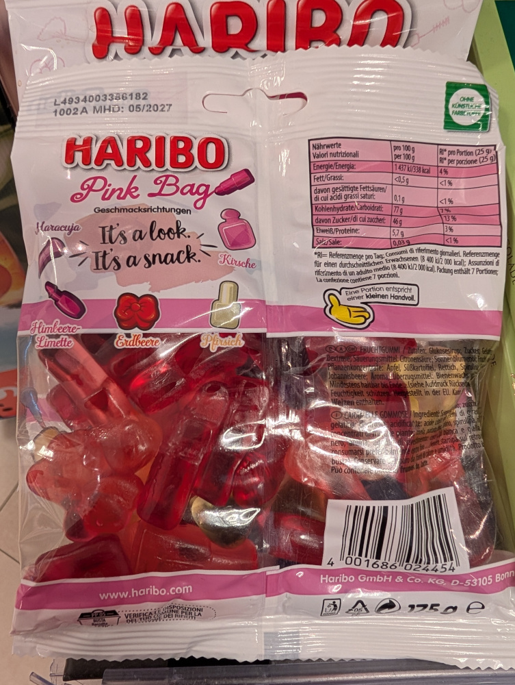 Rückseite einer Packung „HARIBO Pink Bag“ im Verkaufsregal. Die Verpackung ist weiß und rosa gestaltet. Neben der Aufschrift „It’s a look. It’s a snack.“ sind stilisierte Beauty-Produkte abgebildet. Darunter werden die Geschmacksrichtungen Maracuja, Kirsche, Himbeere-Limette, Erdbeere und Pfirsich den verschiedenen Fruchtgummiformen zugeordnet. Zu sehen sind unter anderem ein Flakon, eine Schleife und ein länglicher Kosmetikartikel. Rechts befinden sich Nährwertangaben und Zutatenliste; unten ist als Hersteller die Haribo GmbH & Co. KG, D-53105 Bonn, angegeben.