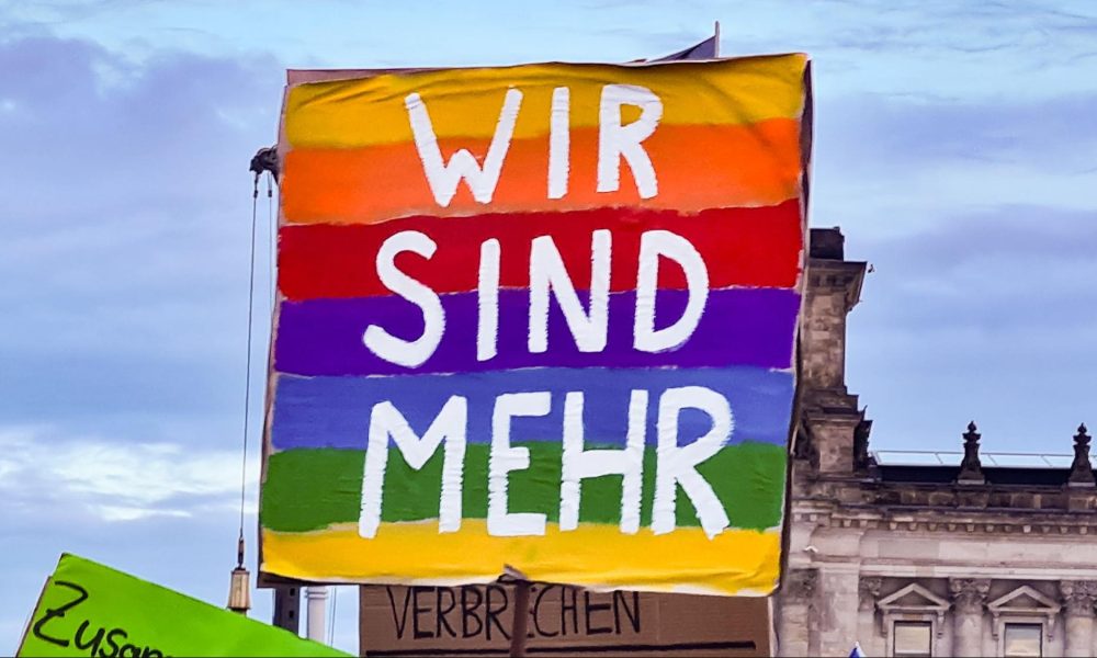 Foto von einem Demoplakat. Das Plakat ist in den Farben des Regenbogens angemalt und darauf steht in weiß WIR SIND MEHR